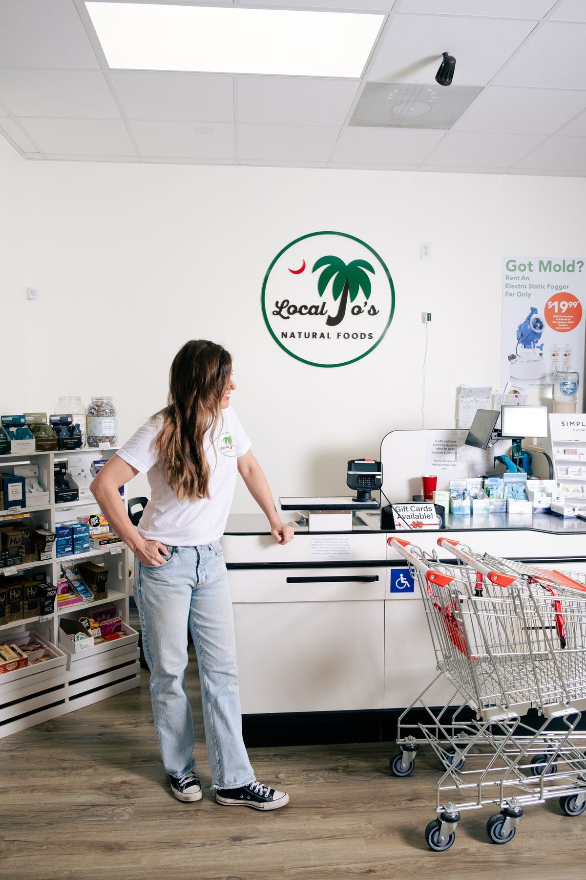 A staff member leaning on the checkout counter under the Local Jo's Natural Foods wall sign, shopping carts beside the register